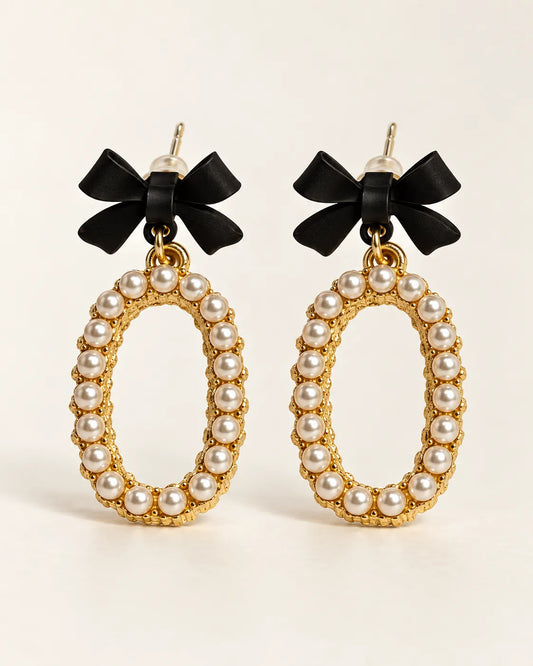 Black bow stud earrings with oval pearl-look bead drops on a cream background