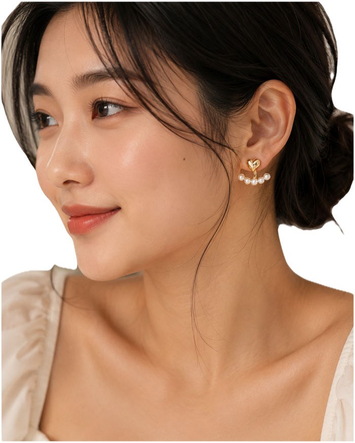 A close-up of a woman wearing a delicate gold heart-shaped dangle earring adorned with small white pearls, shown against a clean white background.