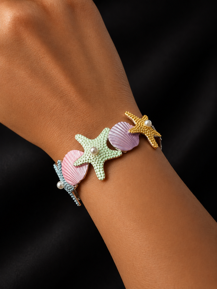 A close-up of a person's wrist wearing a colorful bracelet with starfish and shell motifs in pastel green, pink, lavender, and gold on a black background.