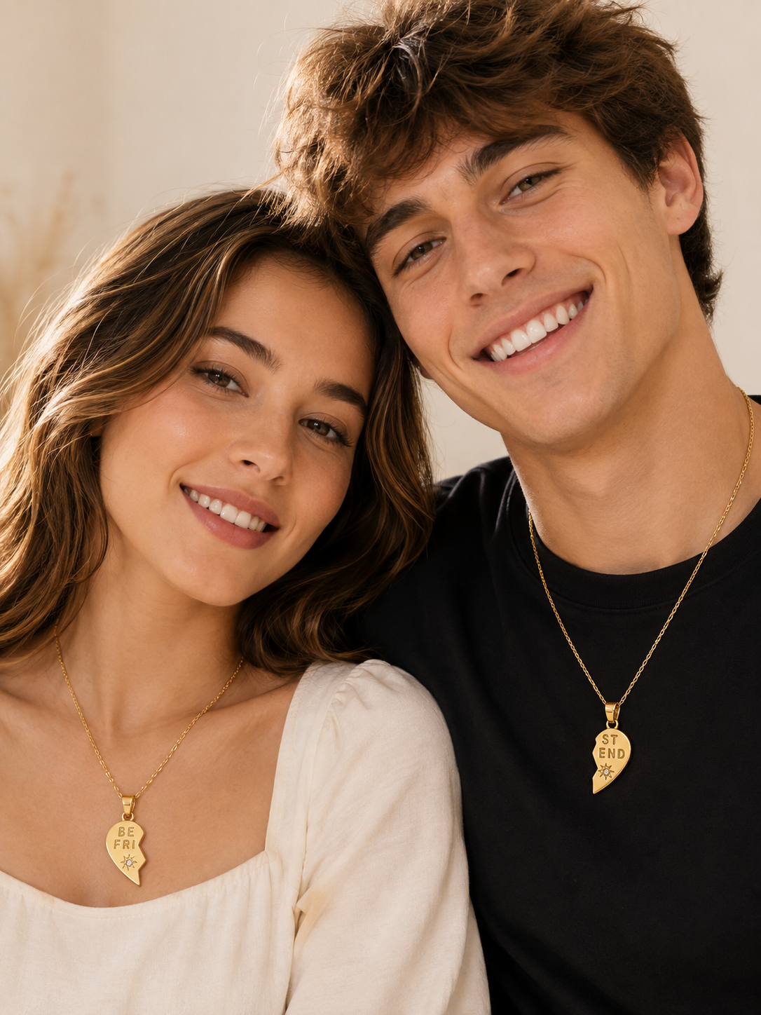 A smiling friend wearing matching gold heart pendant necklaces that form a broken-heart pair, posed closely together against a warm beige background.