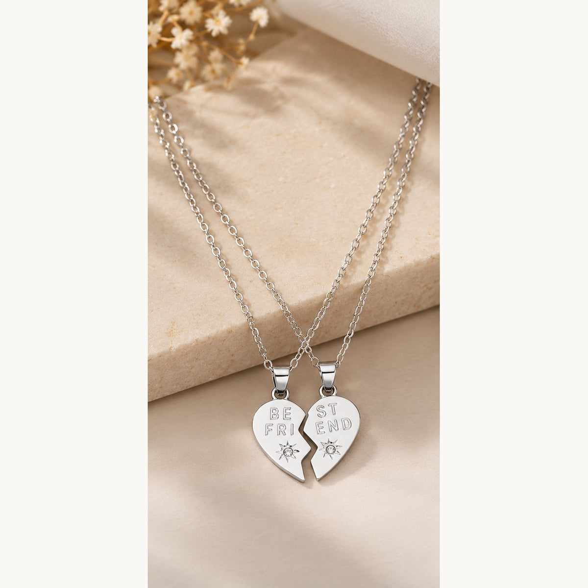 Two silver friendship necklaces with half-heart pendants that fit together, displayed on a beige fabric surface with a small dried floral accent.