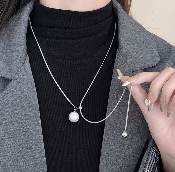 A close-up of a person wearing a delicate silver chain necklace with a white pearl pendant, styled over a black ribbed turtleneck and gray blazer.