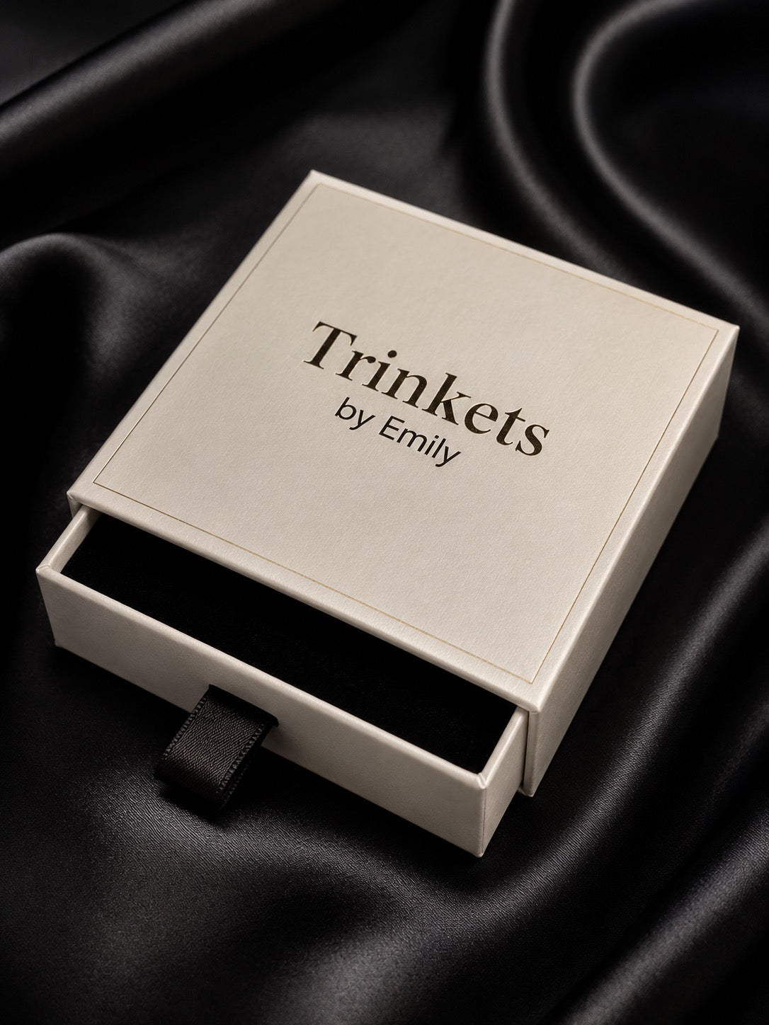 A white-colored jewelry gift box with the brand name 'Trinkets by Emily' printed on the lid, shown with its drawer pulled open to reveal a black interior, set against a draped black satin background.