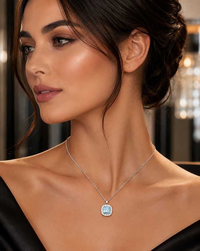 A close-up of a woman wearing a delicate silver chain necklace with a light blue oval pendant, set against a softly blurred indoor background.