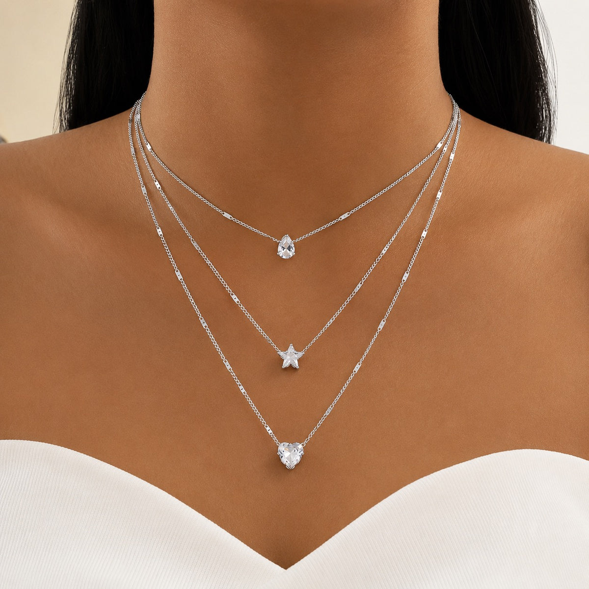 A close-up of a model's neck and upper chest wearing three layered silver necklaces with clear crystal pendants shaped pear, star, and heart.