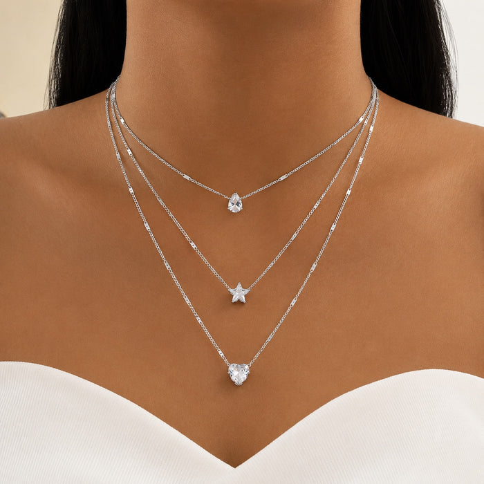 A close-up of a model's neck and upper chest wearing three layered silver necklaces with clear crystal pendants shaped pear, star, and heart.