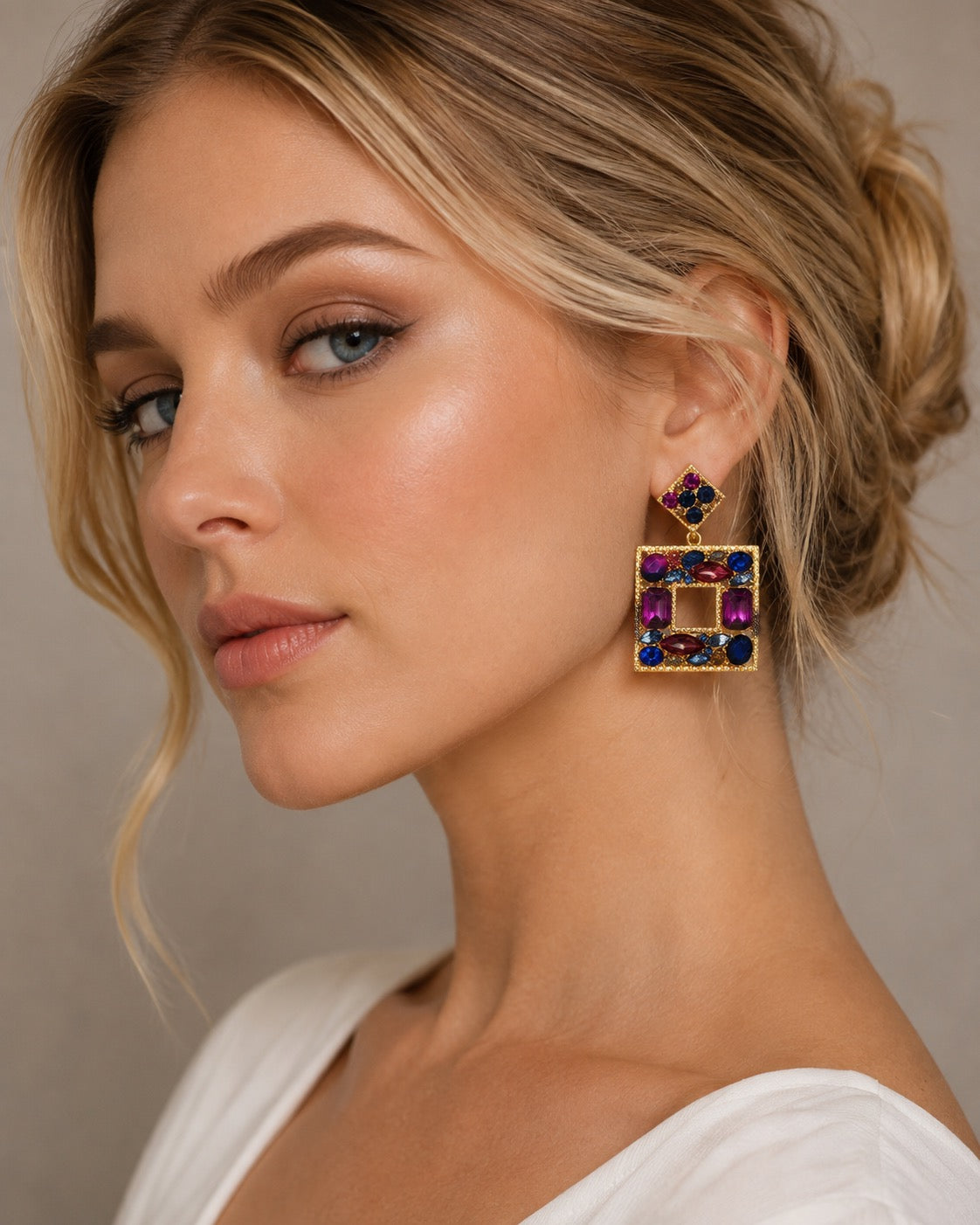 A close-up of a woman wearing a large gold geometric drop earring with colorful gemstones, including purple, blue, and red, against a neutral beige background.