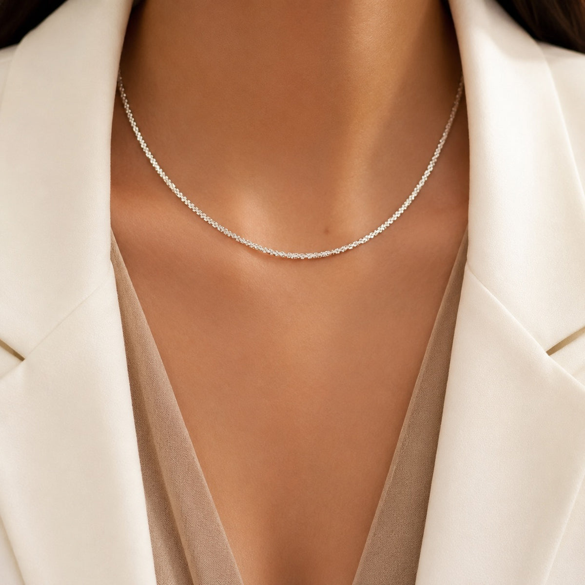 A delicate silver tennis necklace worn by a model, styled with a cream blazer and a beige top.