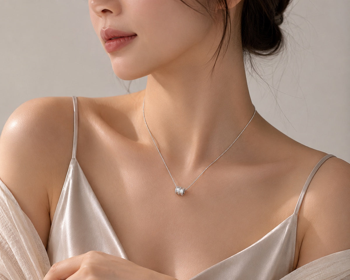 A close-up lifestyle shot of a model wearing a delicate silver necklace with a small, clear gemstone pendant, styled with a satin camisole against a soft neutral background.