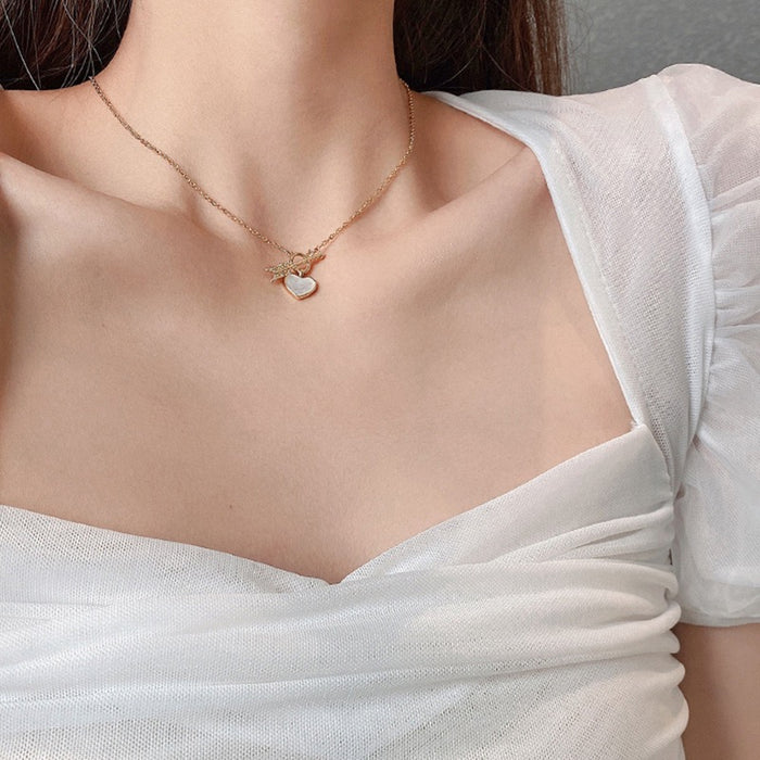 A close-up of a model wearing a delicate gold angel necklace with a small white heart pendant, styled with a white sheer top.