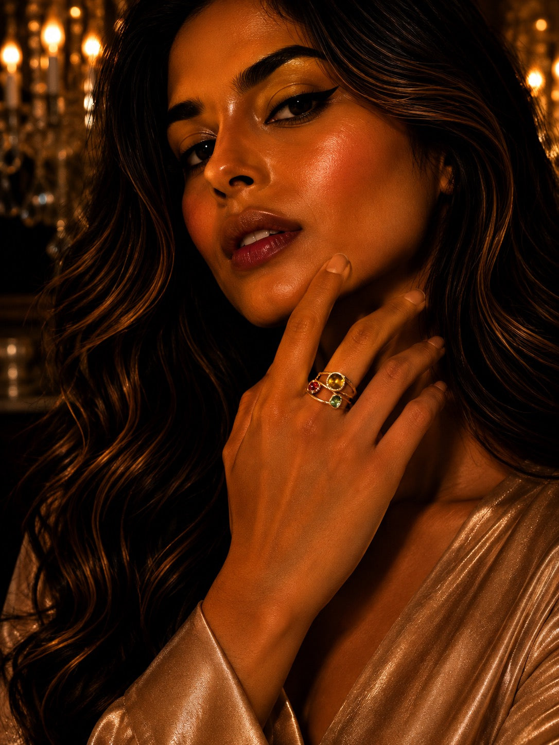 A close-up lifestyle shot of a woman with long wavy brown hair wearing a gold ring with colorful gemstones, set against a warm, dimly lit background with a chandelier.