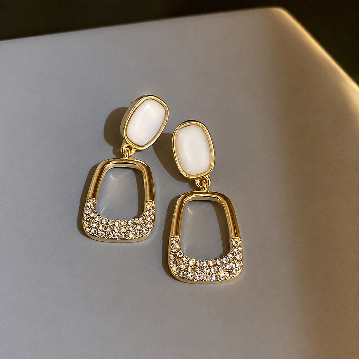 A pair of gold-tone drop earrings with white oval studs and a dangling open arch design set with clear rhinestones, resting on a neutral gray surface.