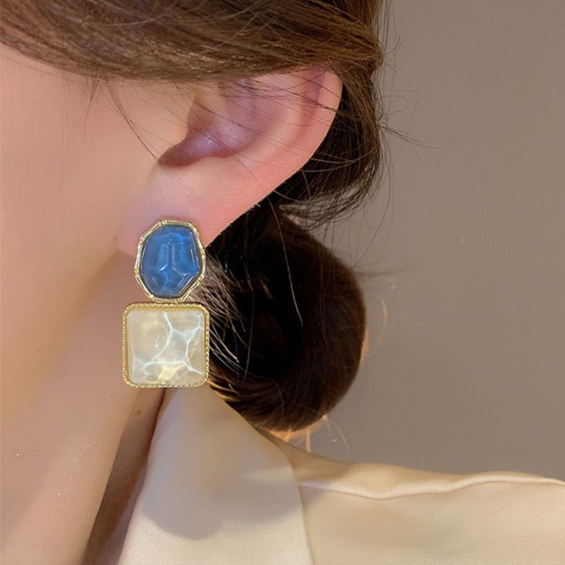 A close-up of a model's ear wearing a two-step drop earring. The top step features a faceted blue stone in a gold bezel, and the bottom step is a square milky white stone in a gold bezel.