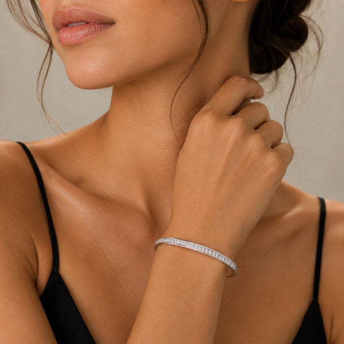 A close-up lifestyle shot of a woman wearing a thin silver tennis bracelet with a continuous line of small clear stones. The image focuses on her neck, hand, and upper chest, showcasing how the bracelet looks on the wrist against a neutral beige background.