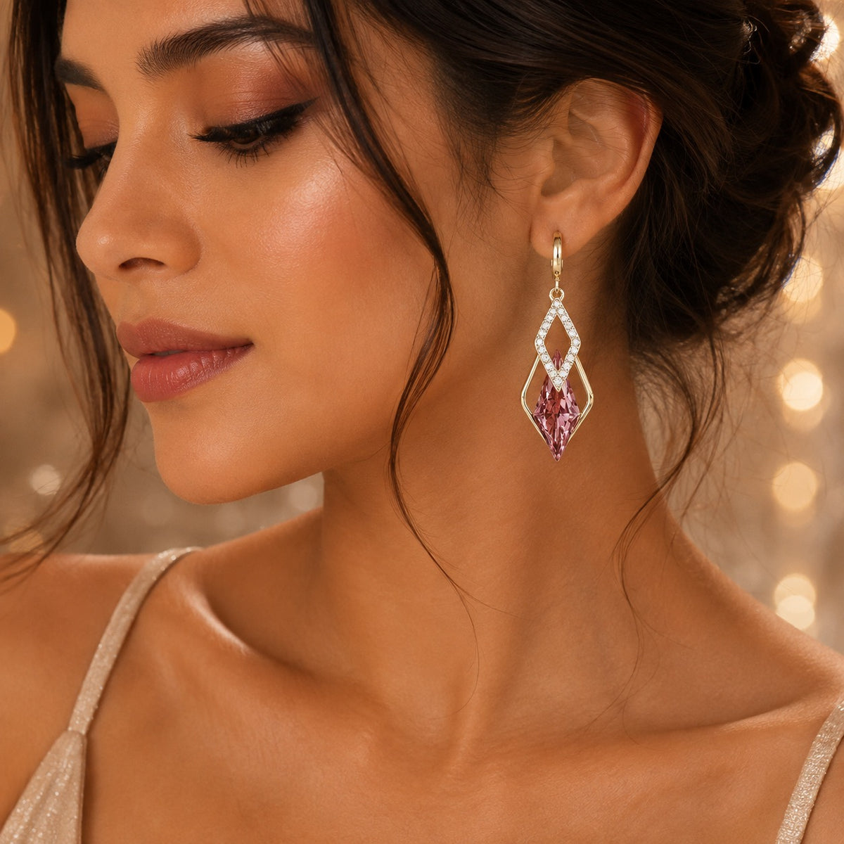 A close-up lifestyle shot of a woman wearing a dangling geometric earring. The earring features a gold-tone frame set with small clear crystals and a central pink gemstone.