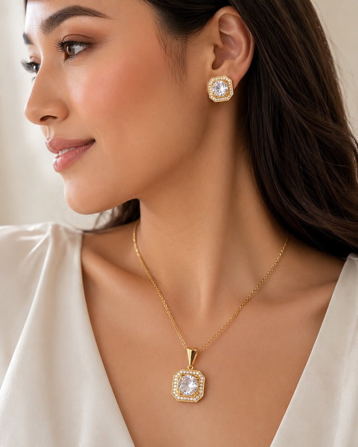 A woman wearing a gold pendant necklace and matching stud earring with clear gemstones, shown in a close-up portrait against a soft neutral background.