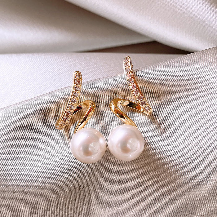 A pair of gold hoop earrings with curved pavé crystal tops and large white freshwater pearls, displayed on a draped beige satin fabric.