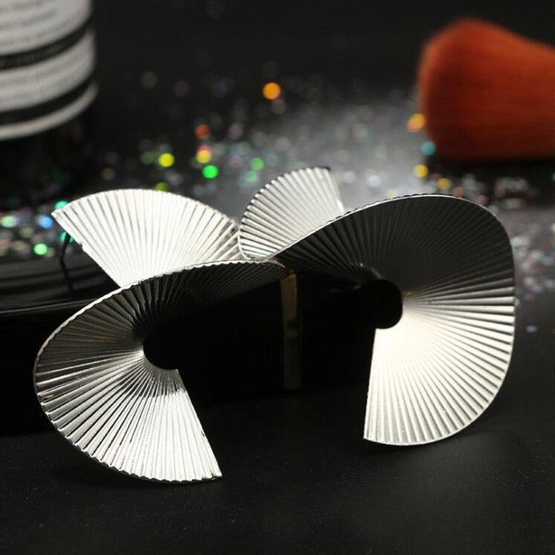 A pair of silver metallic fan-shaped earrings displayed on a black surface with a dark, glittery background and blurred cosmetic props.
