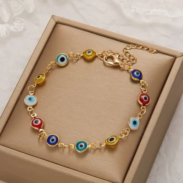 A golden chain bracelet with colorful evil eye charms, displayed inside a beige jewelry box on a white textured fabric background.