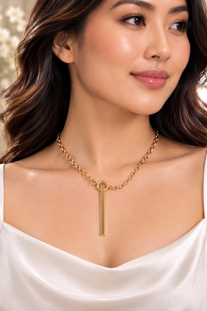 A woman wearing a delicate gold chain necklace with a minimalist vertical bar pendant, styled with a white satin top.