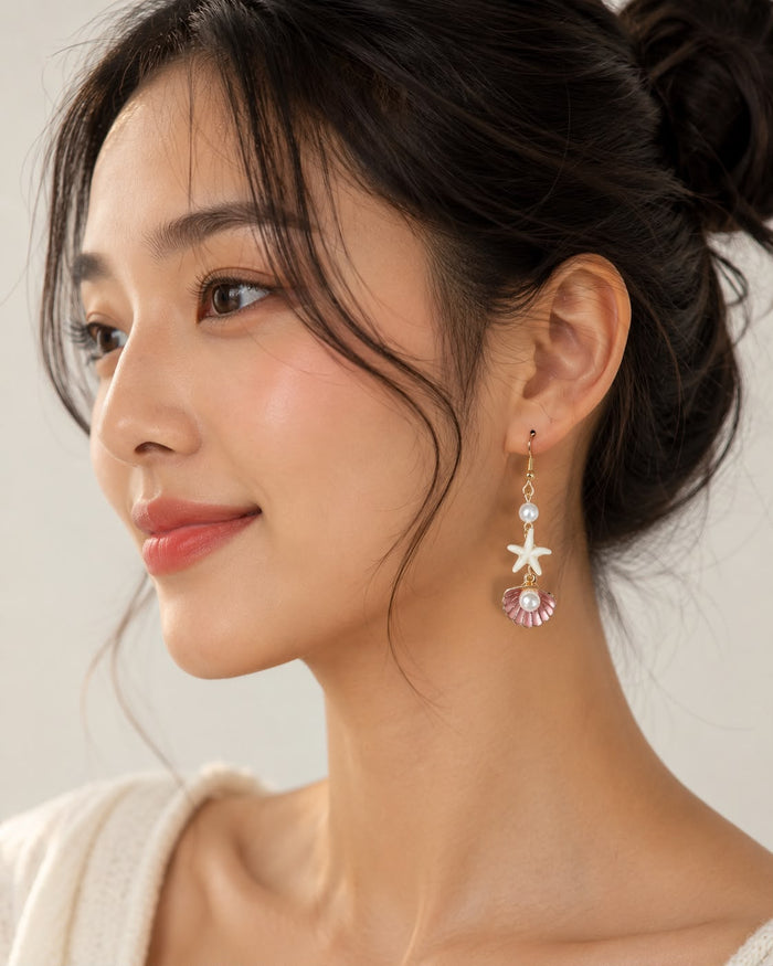A close-up of a woman wearing a dangling earring with a gold starfish, white pearl, and pink shell charm against a soft light gray background.