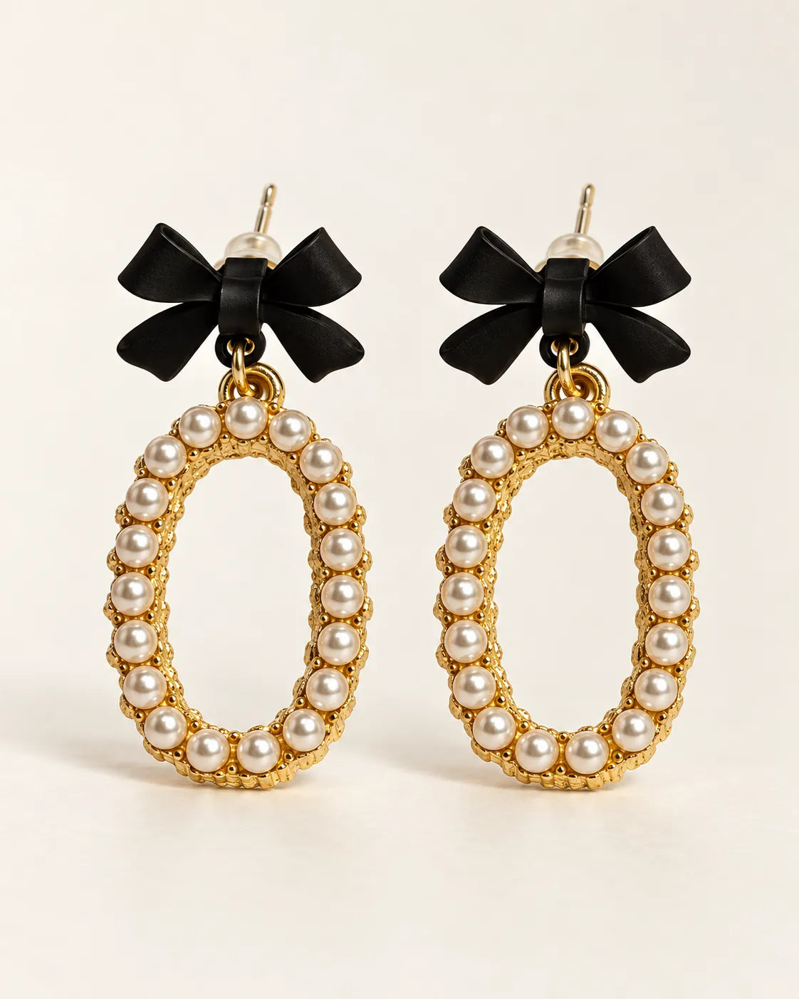 Black bow stud earrings with oval pearl-look bead drops on a cream background