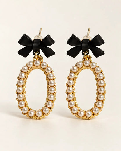 Black bow stud earrings with oval pearl-look bead drops on a cream background