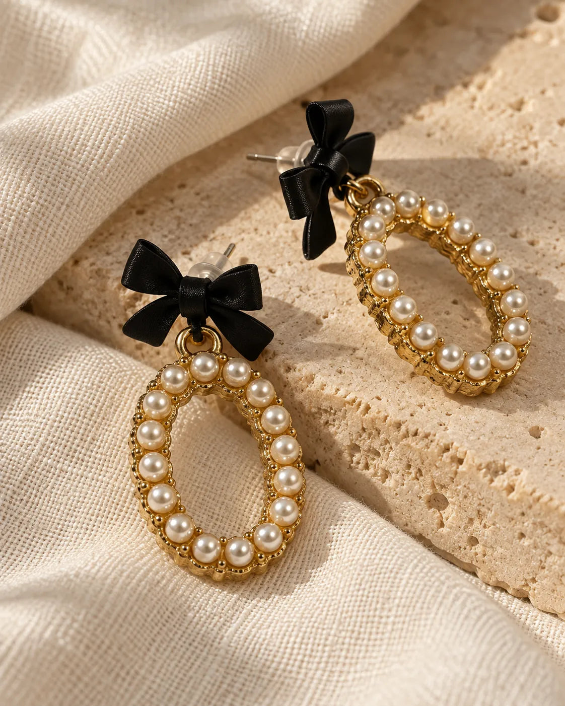 Black bow pearl-look oval earrings resting on stone and cream fabric