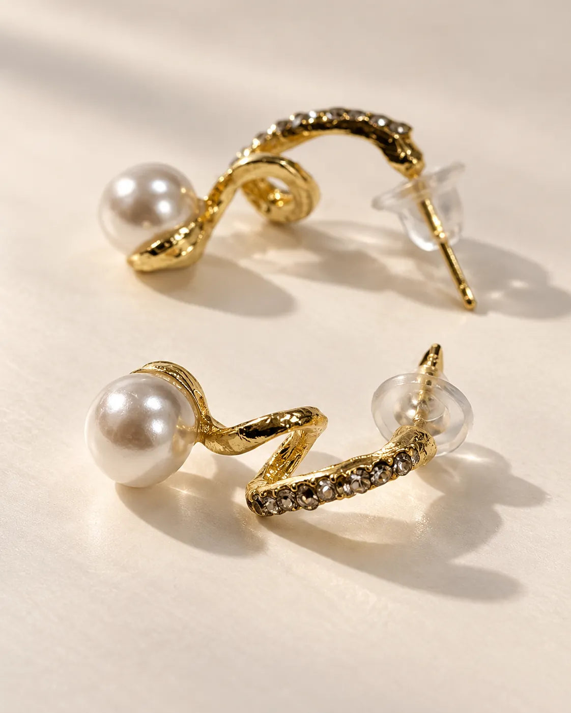 Gold S-shaped rhinestone pearl-look studs on cream showing the clear backings