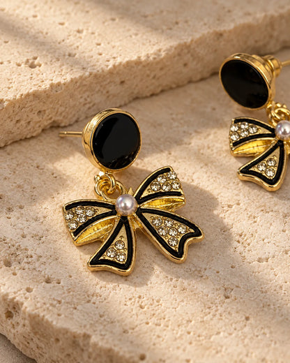 Black and gold-tone bow dangle earrings resting on a stone surface