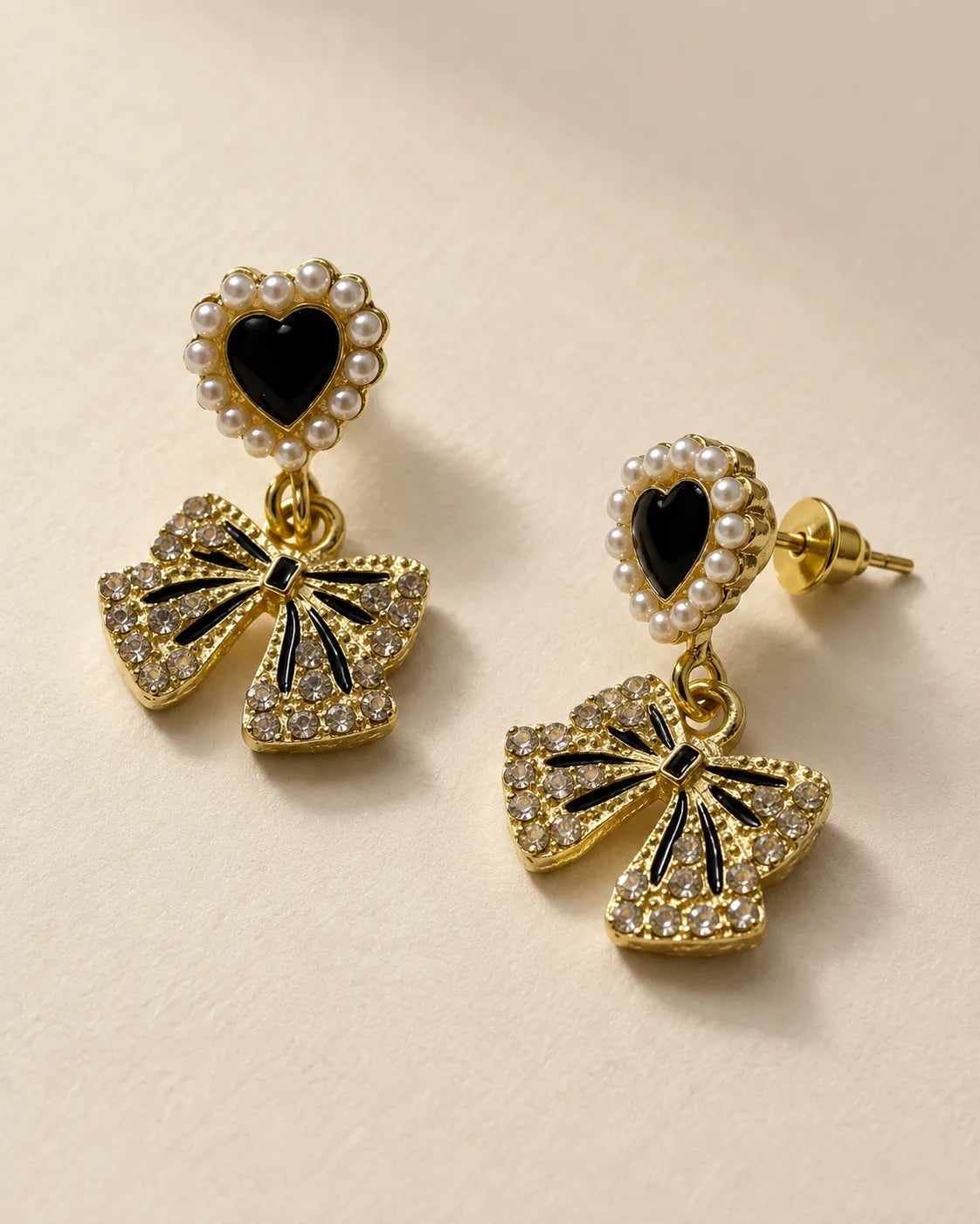 Black enamel heart and gold rhinestone bow dangle earrings on a beige background