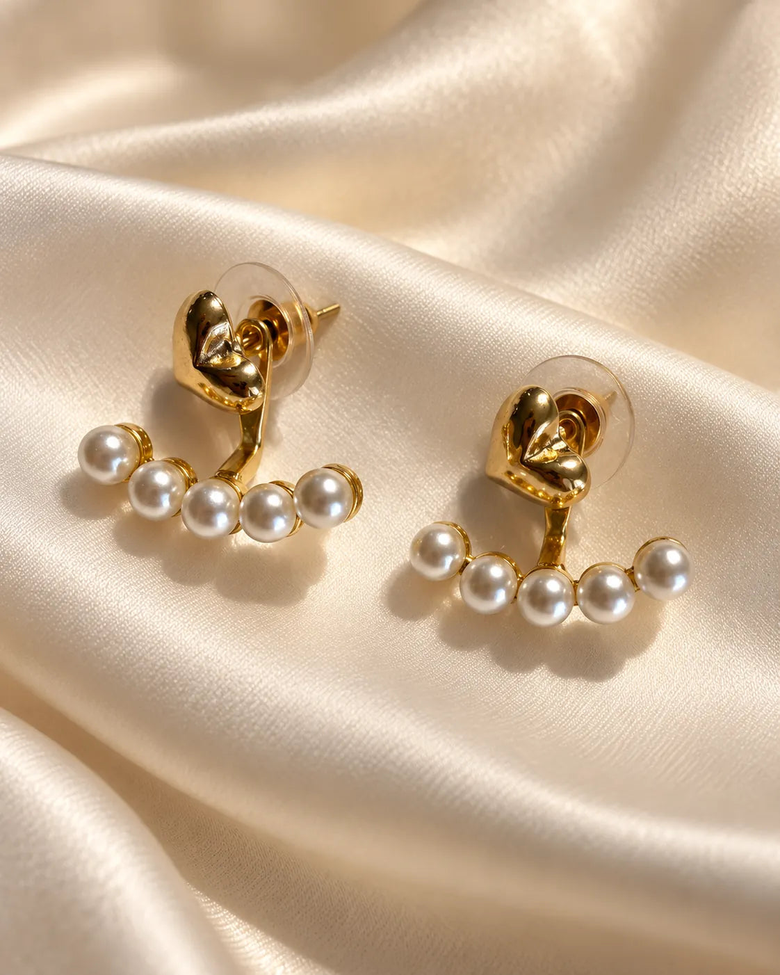 Gold heart pearl-look stud earrings resting on cream satin