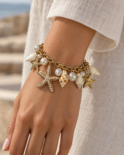 Wrist wearing the gold starfish charm bracelet with a white sleeve by the sea