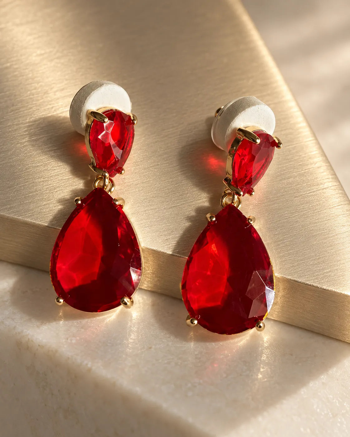 Red two-stone drop earrings on a cream surface in soft sunlight
