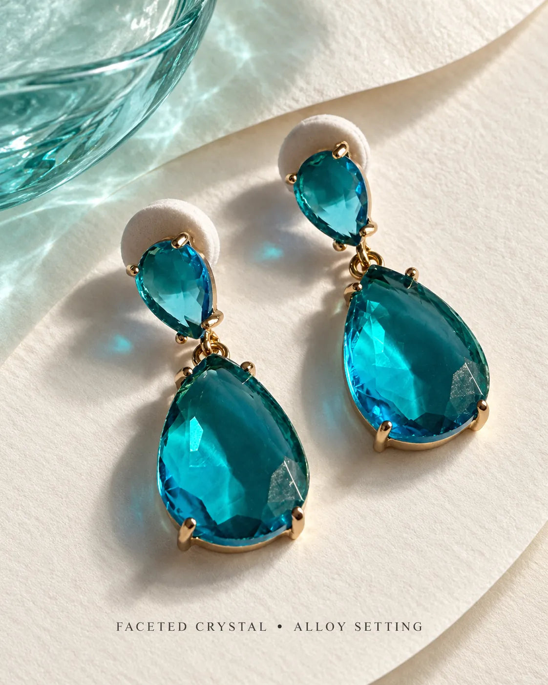Teal-blue crystal teardrop earrings on a pale satin and glass surface