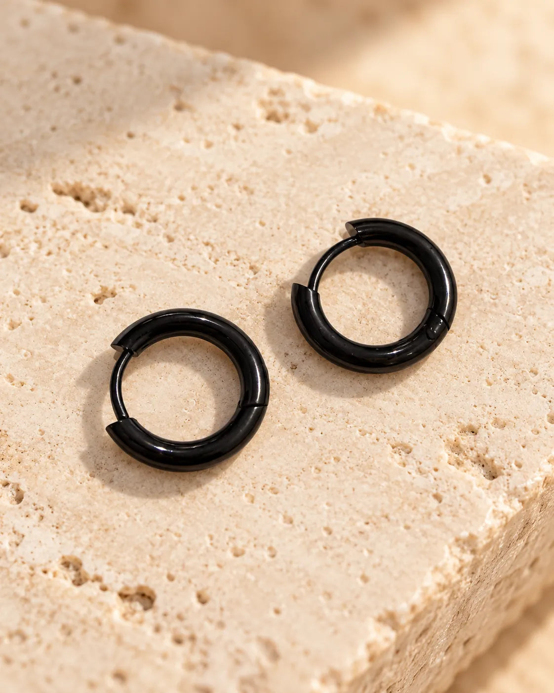 Black stainless steel huggie hoops resting on stone in sunlight