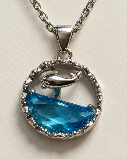 Close-up of the Delphine pendant showing the silver dolphin, blue faceted wave and scalloped frame