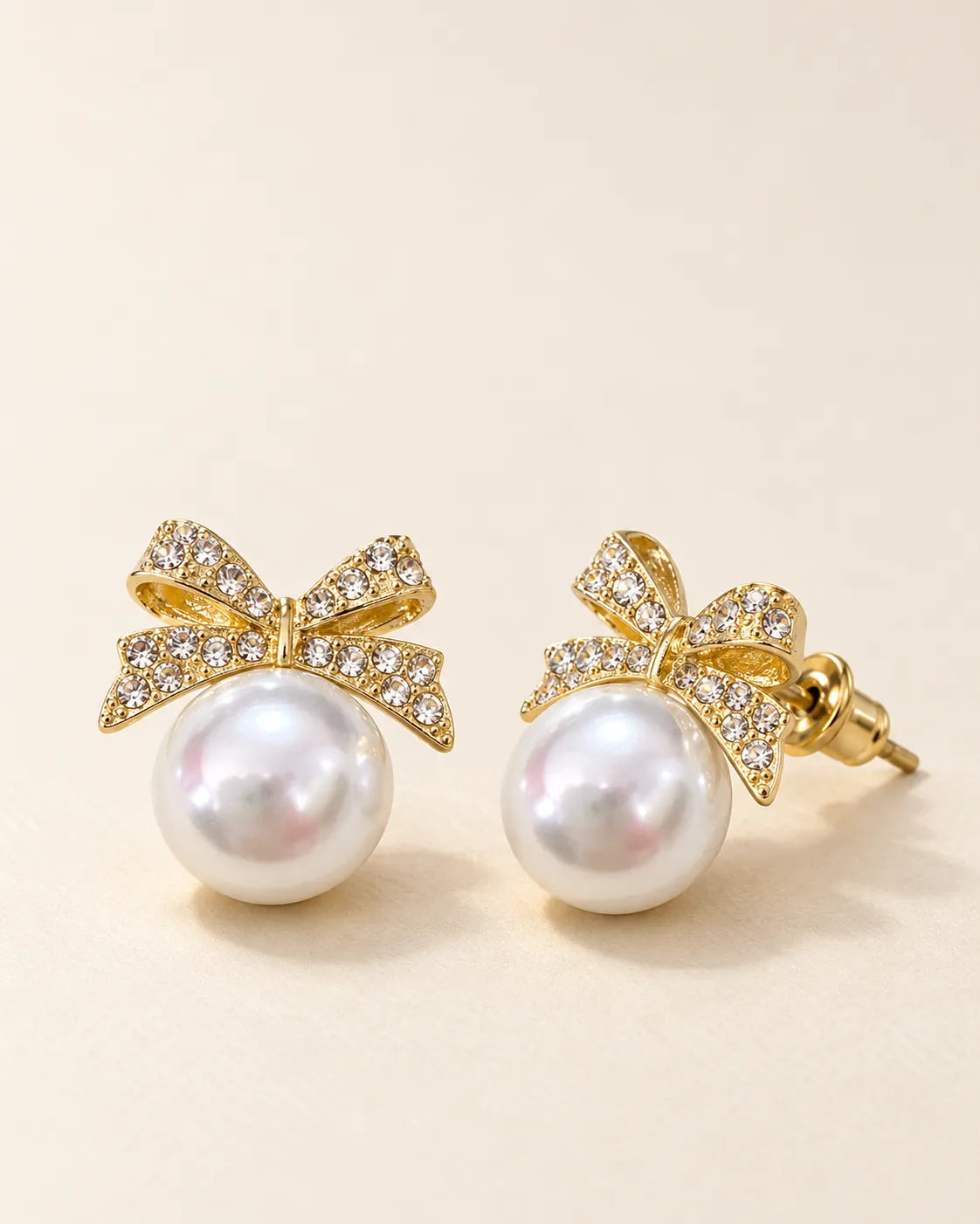 Gold rhinestone bow studs with pearl-look beads on a cream background