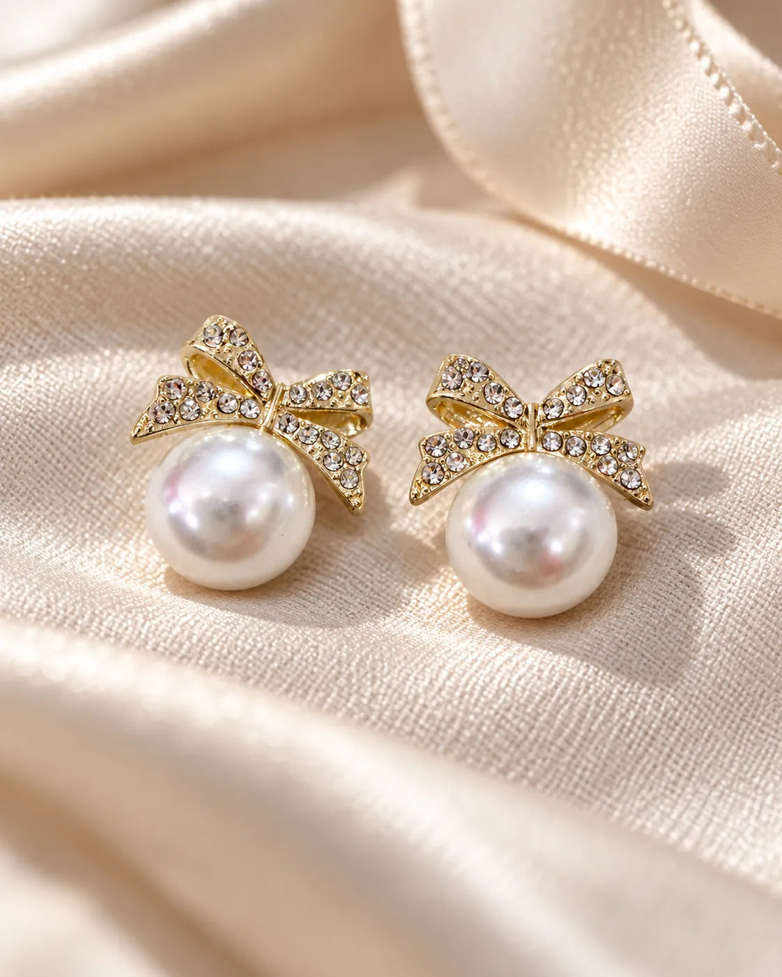 Gold bow and pearl-look stud earrings resting on ivory satin
