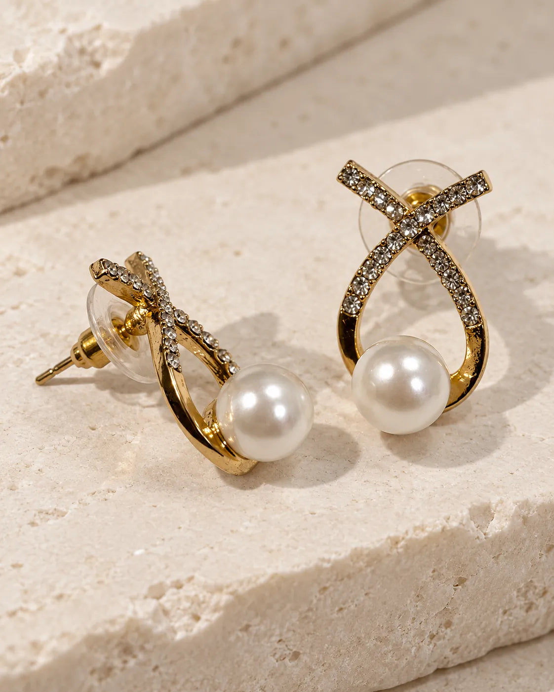 Gold crossed loop stud earrings with pearl-look beads on a stone surface