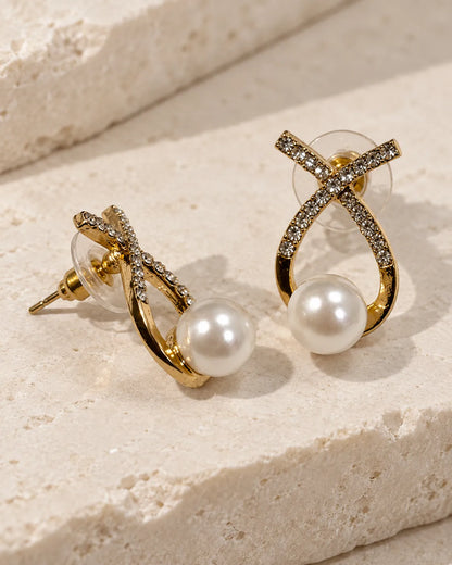 Gold crossed loop stud earrings with pearl-look beads on a stone surface