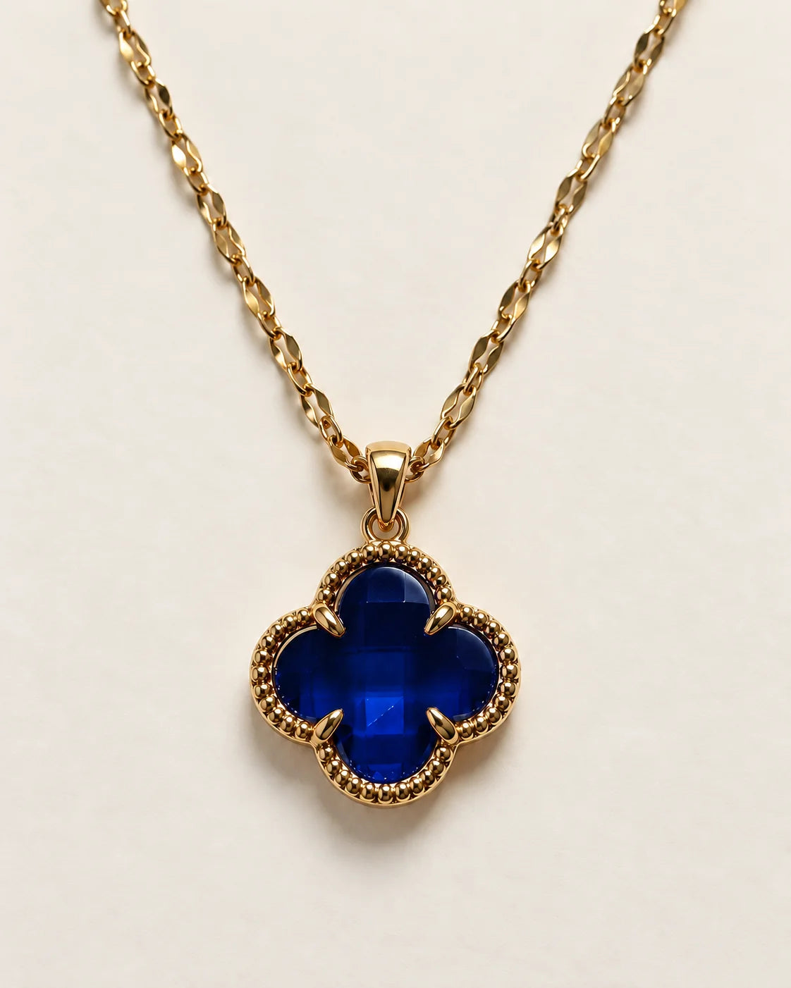 Fortuna sapphire blue clover pendant with a beaded gold-tone border on a cream background