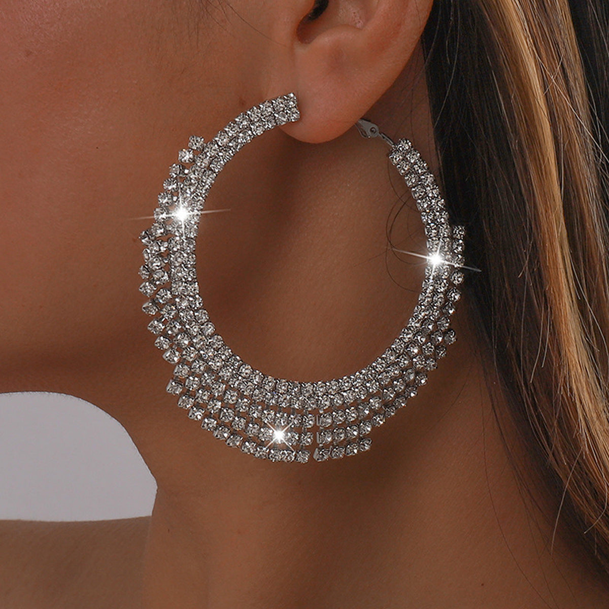 A close-up of a model wearing a large, multi-row silver hoop earring embellished with sparkling clear rhinestones.