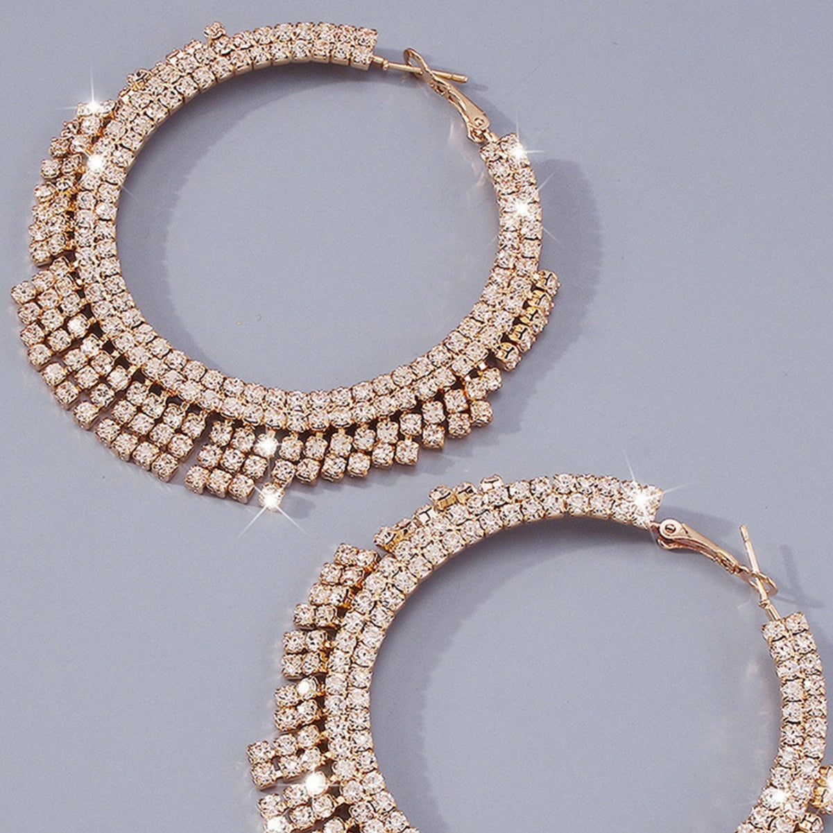 A pair of large gold hoop earrings embellished with multiple rows of sparkling clear rhinestones, displayed on a smooth gray background.