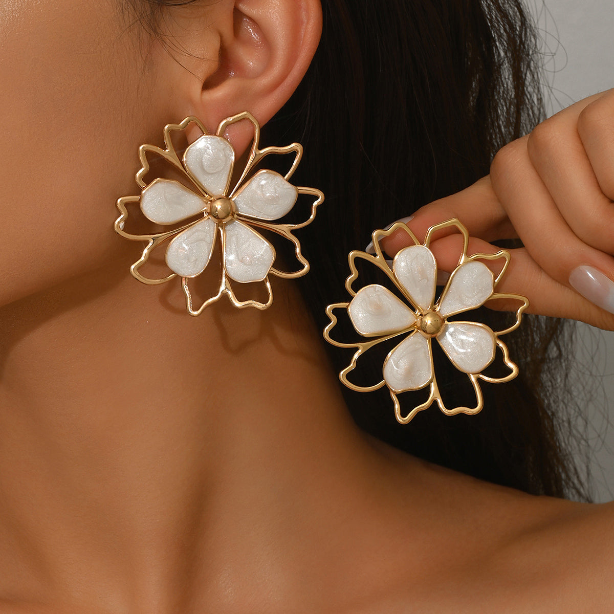 A close-up of a person wearing large flower-shaped earrings with white mother-of-pearl petals and gold-tone metal outlines, while holding the matching earring against a neutral background.