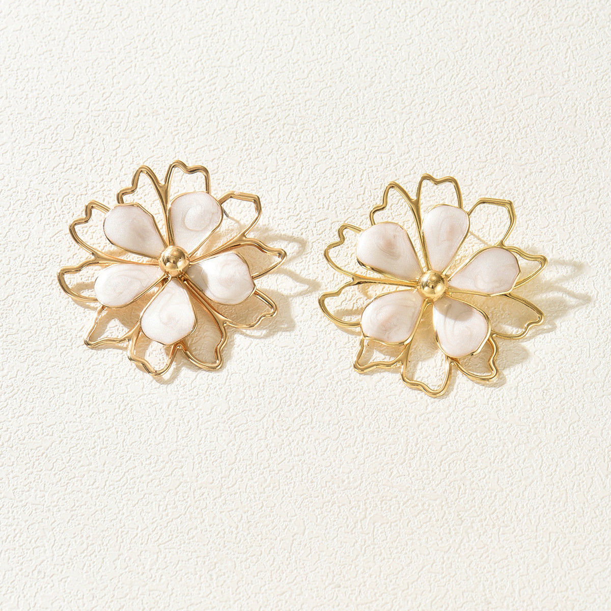 A pair of gold flower-shaped stud earrings with white mother-of-pearl petals, displayed on a textured off-white background.