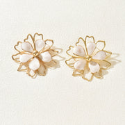 A pair of gold flower-shaped stud earrings with white mother-of-pearl petals, displayed on a textured off-white background.