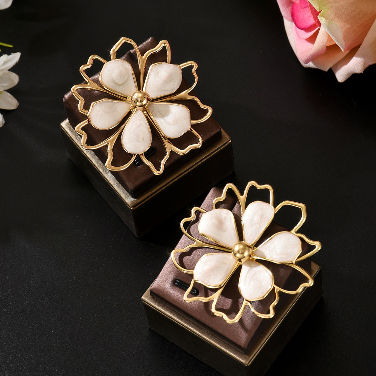 Two white flower-shaped ear rings with gold-tone metal outlines and centers, displayed on dark brown presentation boxes against a black background with soft floral props.