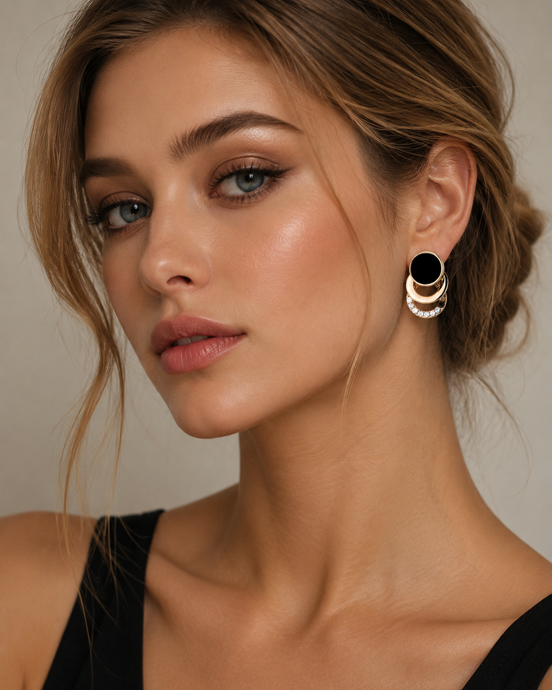 A close-up of a woman wearing a pair of gold-toned geometric drop earrings with black circular centers and small clear stones, against a soft beige background.
