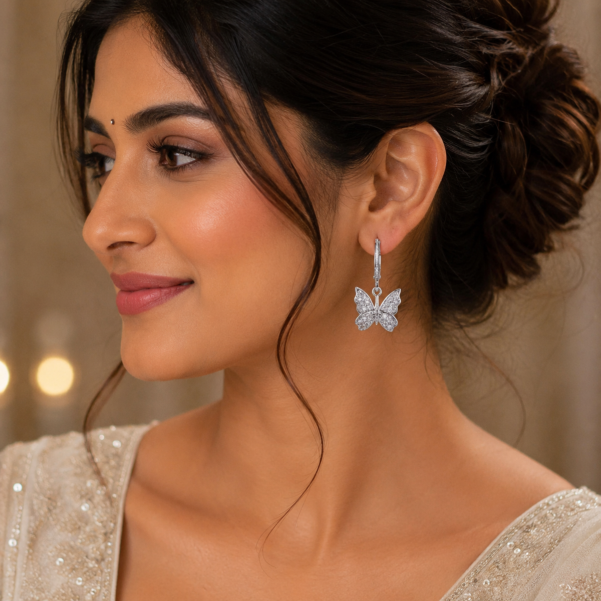 A woman in profile wearing a silver butterfly-shaped drop earring adorned with sparkling clear stones, set against a softly blurred warm-toned background.
